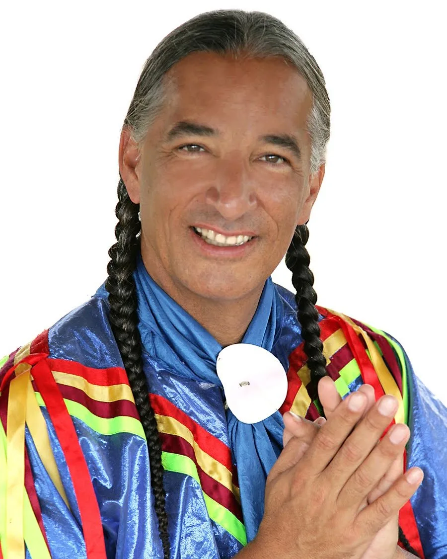 Kevin Locke smiling with hands pressed together, long gray braids, wearing a shiny blue ribbon shirt with red, yellow and green ribbons and a white shell at his collar.