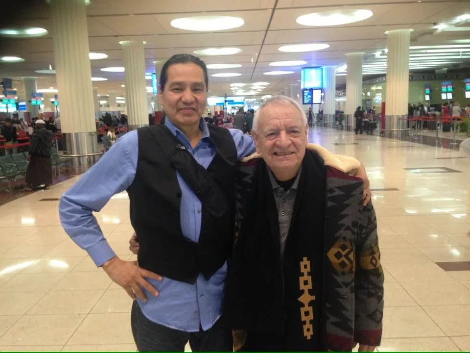 Two smiling men pose arm in arm in a busy airport terminal; one wears a blue shirt and black vest, the other an older man in a patterned blanket coat.