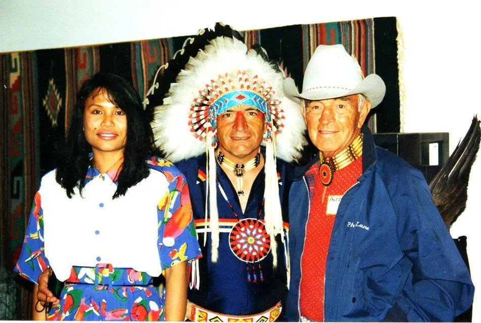Three people smile in front of a woven blanket: a woman in a colorful floral dress, Phil Lane Jr. in a feathered headdress and beaded regalia, and his father in a white cowboy hat and blue jacket.