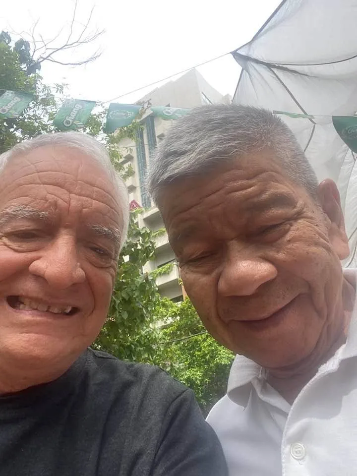 Two smiling older men with gray hair pose close together for a selfie outdoors, one in a dark shirt and one in a white collared shirt, with trees, green banners and a building behind them.