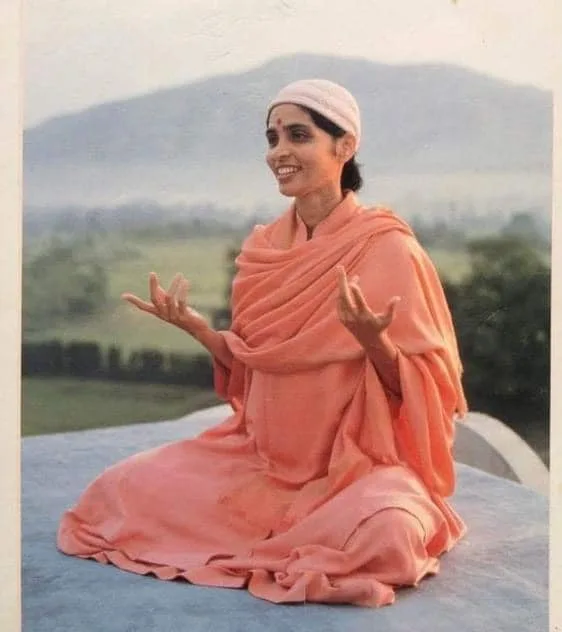 Gurumayi Chidvilasananda, smiling and wearing flowing orange robes and a white cap, sits cross-legged with hands raised, green hills and a mountain behind her.