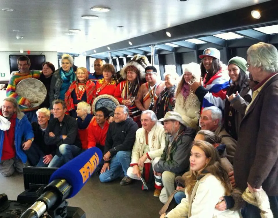 A large group of Indigenous and non-Indigenous people pose together indoors, some in feather headdresses and regalia, holding hand drums, with a BFMTV microphone in the foreground.