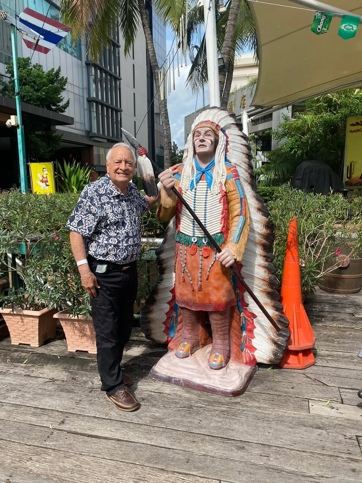 Phil Lane Jr., smiling in a patterned shirt, stands with his hand on the shoulder of a six-foot painted statue of a chief in a feathered headdress outside a hotel in Southeast Asia.