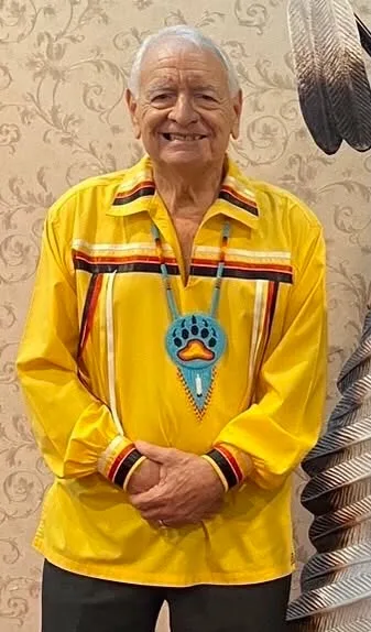 A smiling elder with white hair wearing a yellow ribbon shirt with red and black trim and a beaded turquoise bear paw medallion, standing before patterned wallpaper beside large feathers.