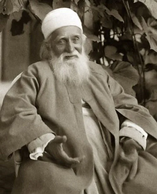 Sepia portrait of 'Abdu'l-Bahá, an elderly man with a long white beard and white head covering, seated in a robe before leafy vines, one hand extended.