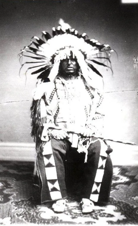Historic black-and-white portrait of a seated Dakota man in a full eagle-feather headdress, beaded buckskin shirt and leggings, holding a long pipe across his lap.
