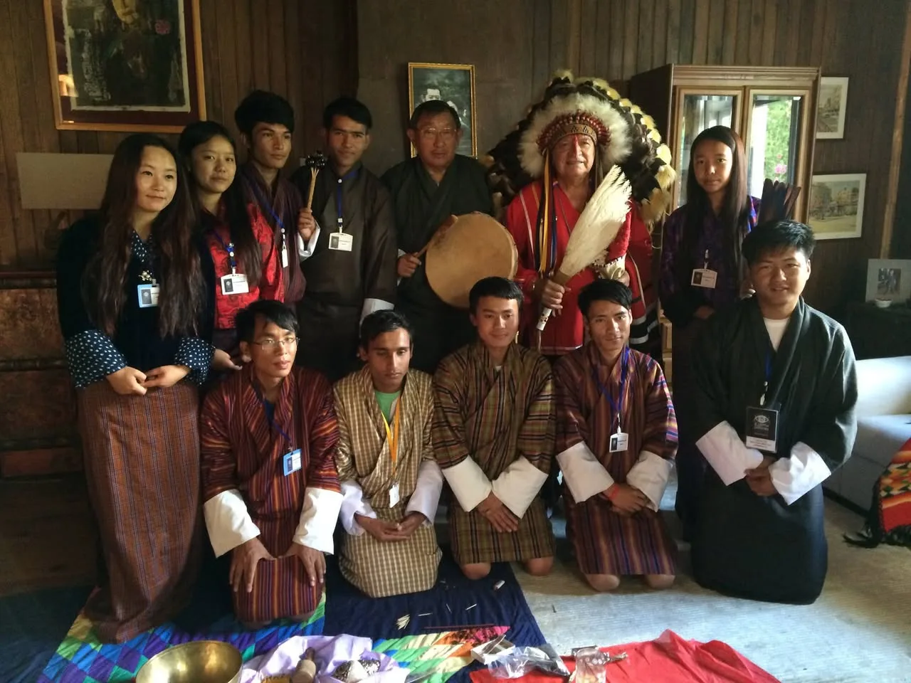 A group of young Bhutanese people in traditional gho and kira, wearing lanyards, pose indoors with an older man holding a drum and an elder in a feathered headdress holding a white feather fan.