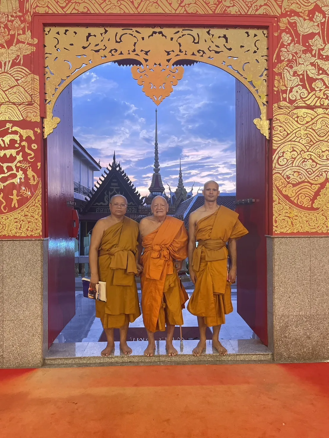 Phil Lane Jr., the Abbot of Wat That Temple and Tonska Robert stand barefoot in saffron robes in an ornate red and gold temple doorway, with spires and a dawn sky behind.