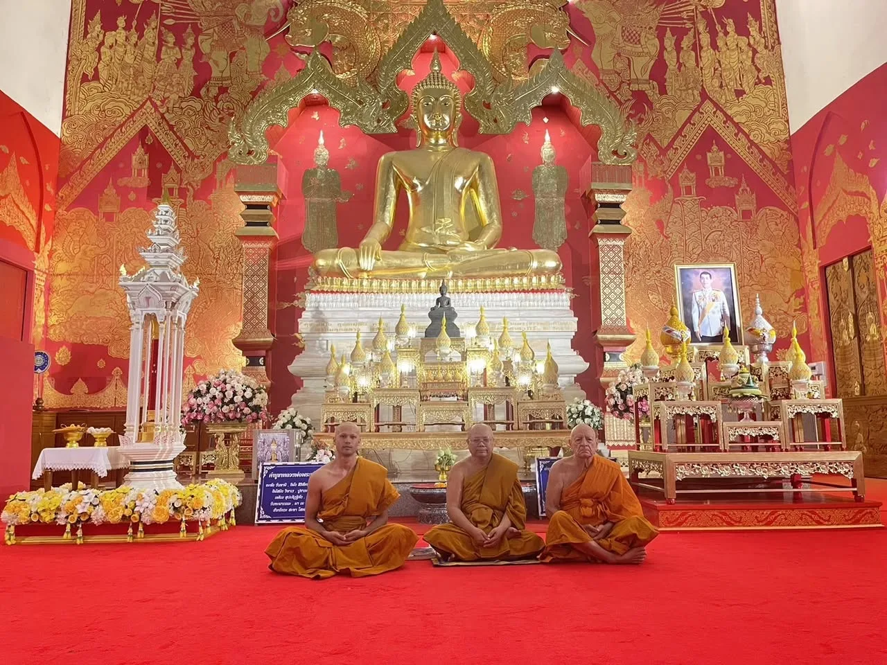 Three monks in saffron robes sit cross-legged on a red carpet before a large golden seated Buddha in an ornate red and gold temple hall decorated with flowers.