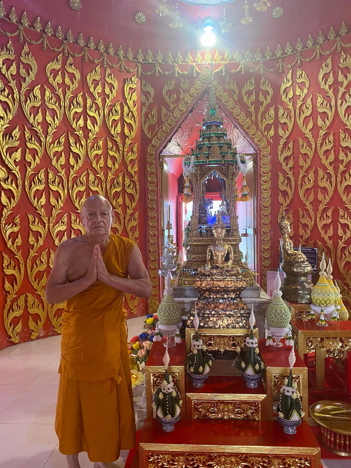 An elder in saffron monk's robes stands with palms pressed together before a gold-leafed seated Buddha and ornate shrine, surrounded by red walls with gold flame patterns.