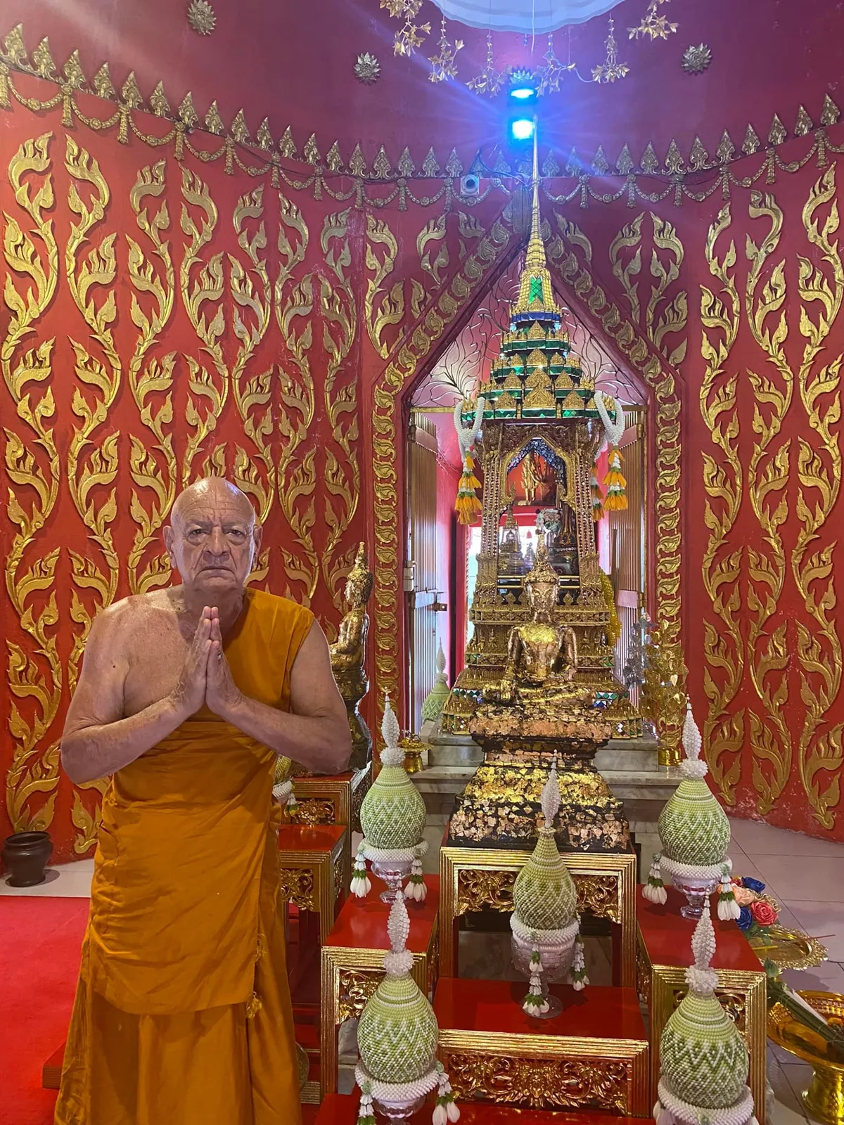 The same robed elder, hands joined in prayer, beside an ancient gilded Buddha on a tiered altar adorned with green beaded offerings and flower garlands in a red and gold shrine.