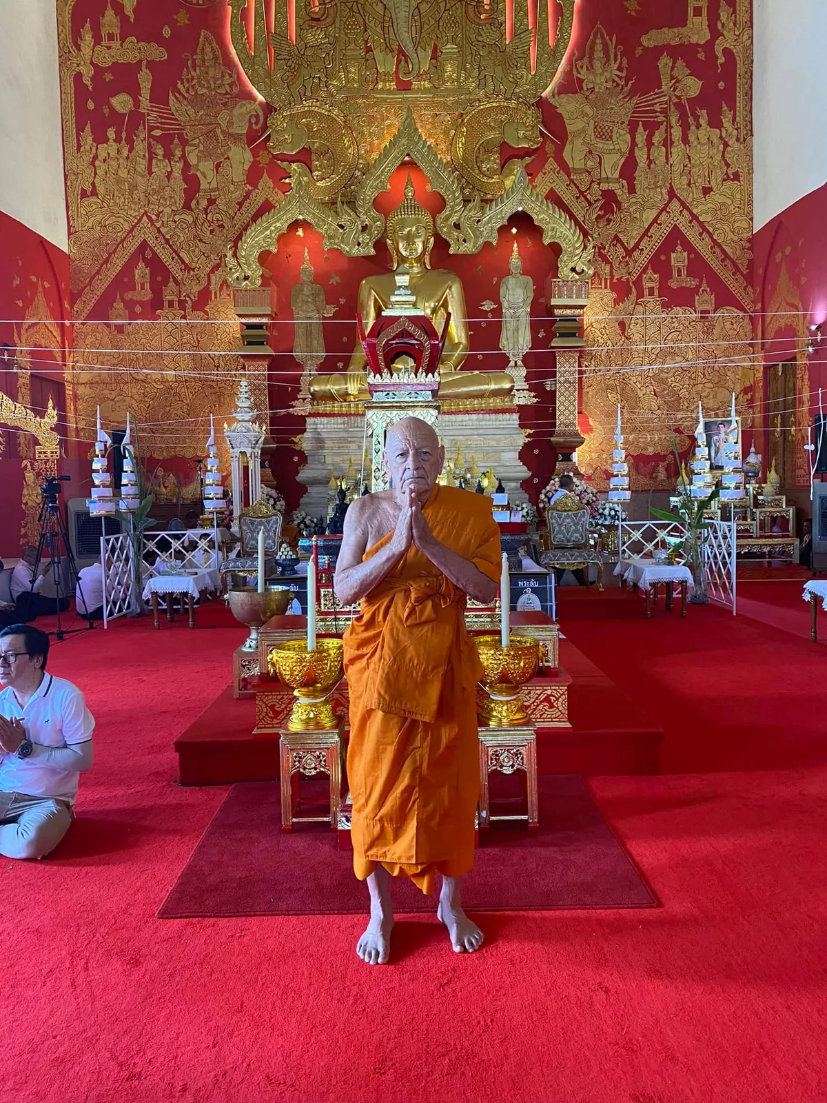 An elderly monk in saffron robes stands barefoot with palms pressed together on a red carpet before a golden seated Buddha in an ornate red and gold temple hall.