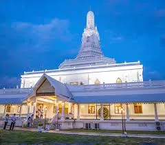 A large white Buddhist temple building with a tall tiered spire, lit up at dusk against a deep blue sky, with a few people walking near the entrance.