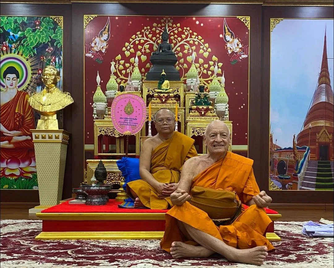 Two Buddhist monks in saffron robes sit in a Thai temple hall; the smiling elder in front holds an alms bowl in his lap, before a golden altar with Buddha statues.