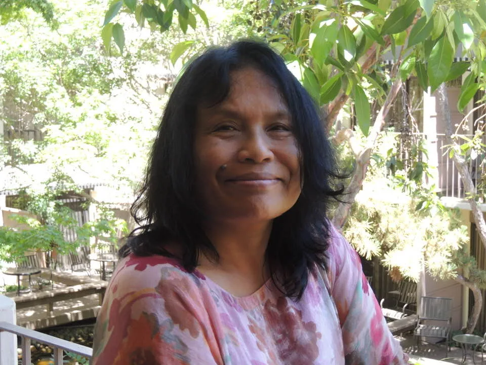 A smiling woman with shoulder-length black hair and a pink floral blouse sits outdoors on a sunny balcony, with green trees and a courtyard behind her.
