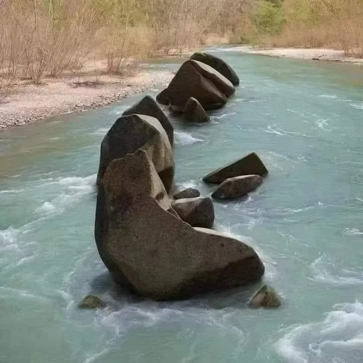 Large boulders in a turquoise river with white rapids, arranged so that at a glance they resemble a seated figure reading a book, beside a pebbled shore and bare shrubs.
