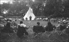 Fotografía en blanco y negro de un grupo grande sentado en sillas plegables formando un círculo sobre el pasto, frente a un tipi rodeado de árboles, con una persona de pie al centro.