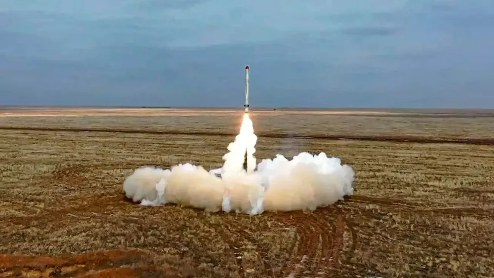 A missile launches upward from a flat, open field, trailing flame and a wide ring of white smoke beneath an overcast sky.