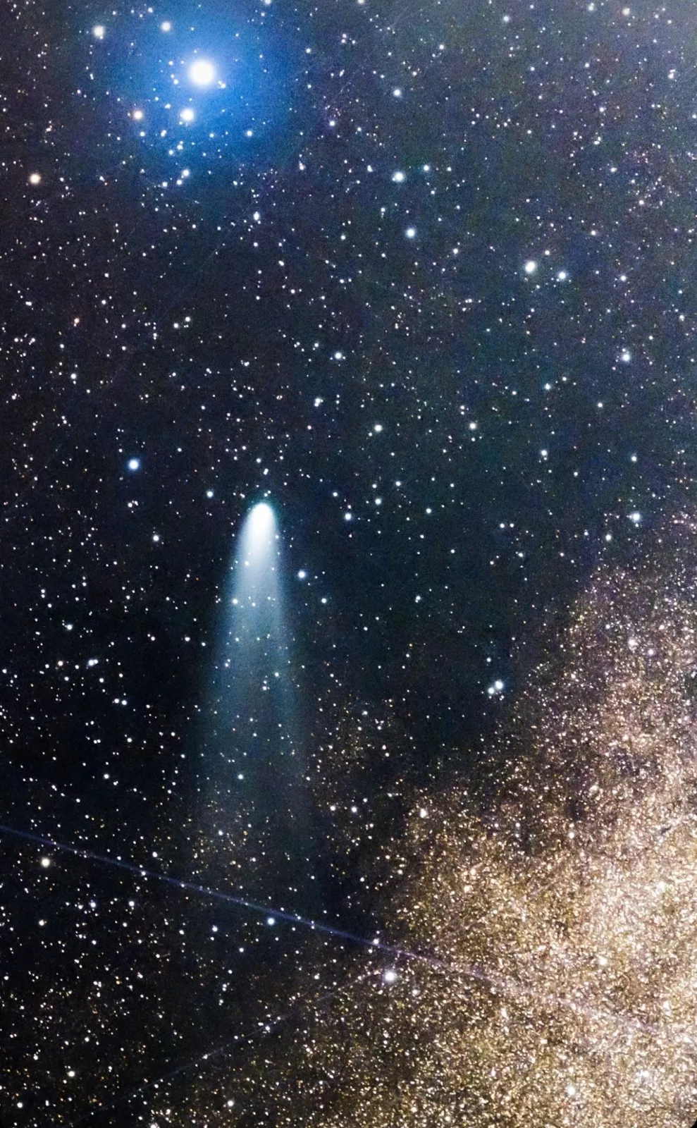 A comet with a glowing blue-green head and long fanning tail streaks across a dense starfield, with a bright blue star cluster above and the golden Milky Way at lower right.