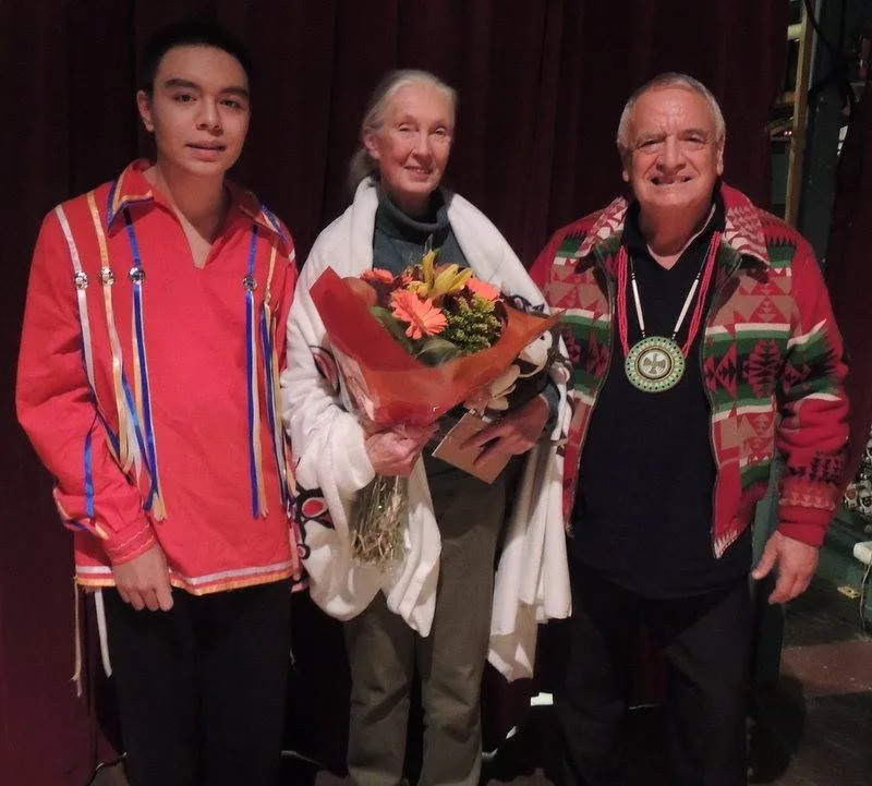 Jane Goodall, holding a bouquet of orange flowers and wearing a white shawl, stands between a young man in a red ribbon shirt and an elder in a patterned jacket and beaded medallion.
