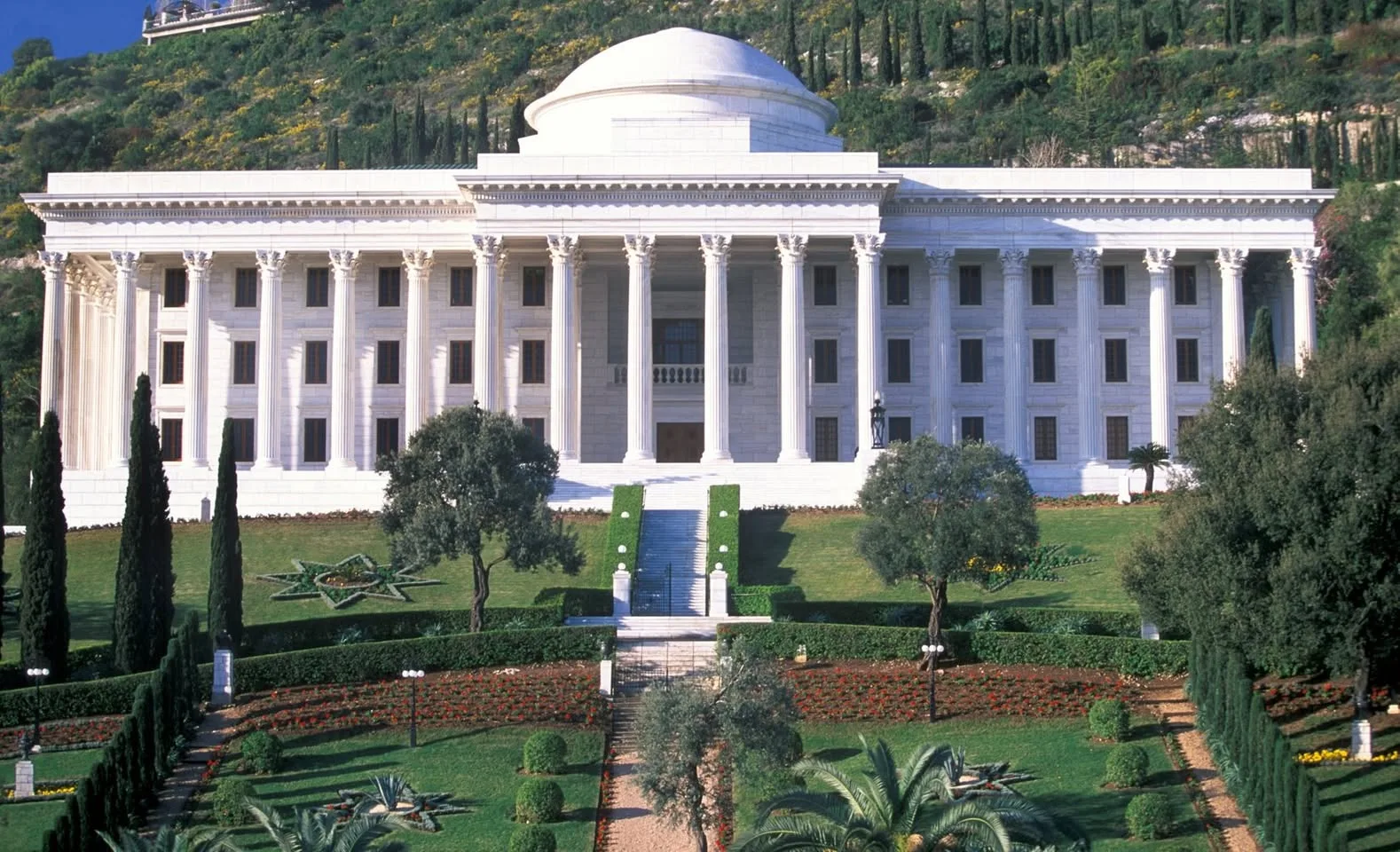 A white marble neoclassical building with tall Corinthian columns and a domed roof stands on a hillside above terraced gardens, cypress trees and a central staircase.