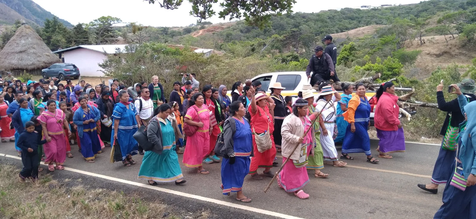 Una multitud de mujeres indígenas con vestidos tradicionales de colores vivos marcha por una carretera de montaña; una sostiene un bastón y al fondo se ven una choza de paja y una camioneta.