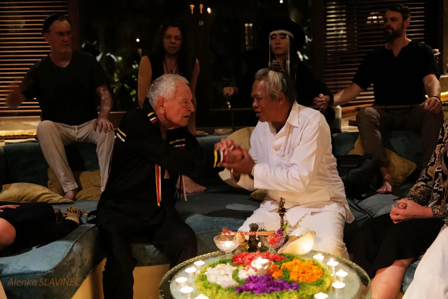 A white-haired elder in a black ribbon shirt clasps hands with a man in white robes beside a candlelit table of colorful flower petals, as others sit around them at night.