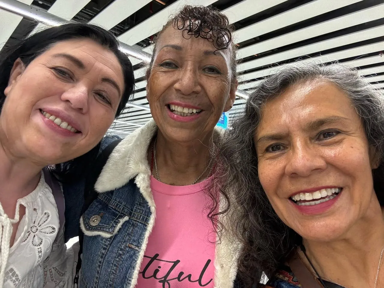 Three smiling women pose close together for a selfie under a striped ceiling; the woman in the middle wears a denim jacket with a fleece collar over a pink shirt.
