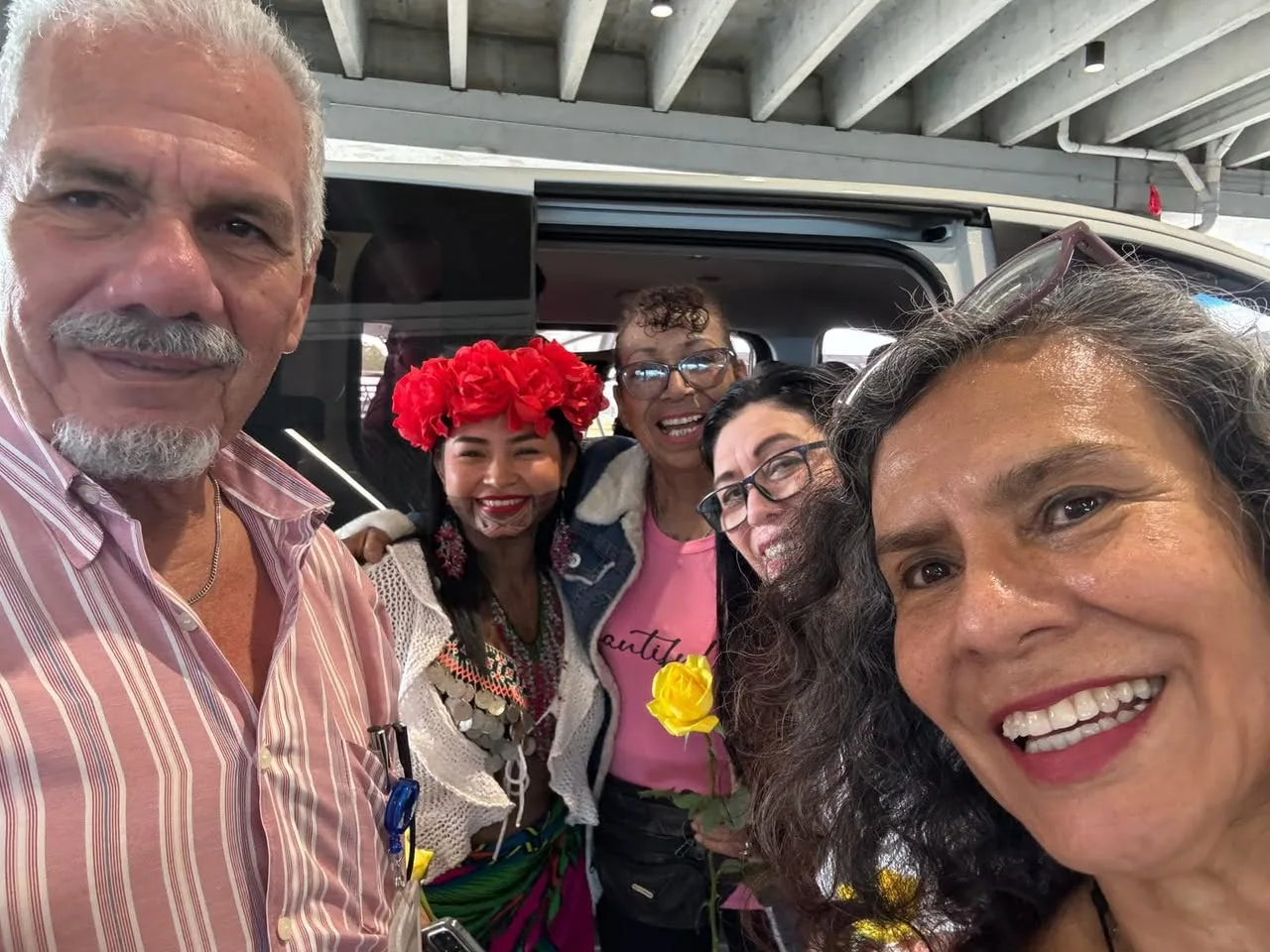 A gray-haired man in a striped shirt and four women pose beside a van; one woman wears a red flower crown and traditional dress, another holds a yellow rose.
