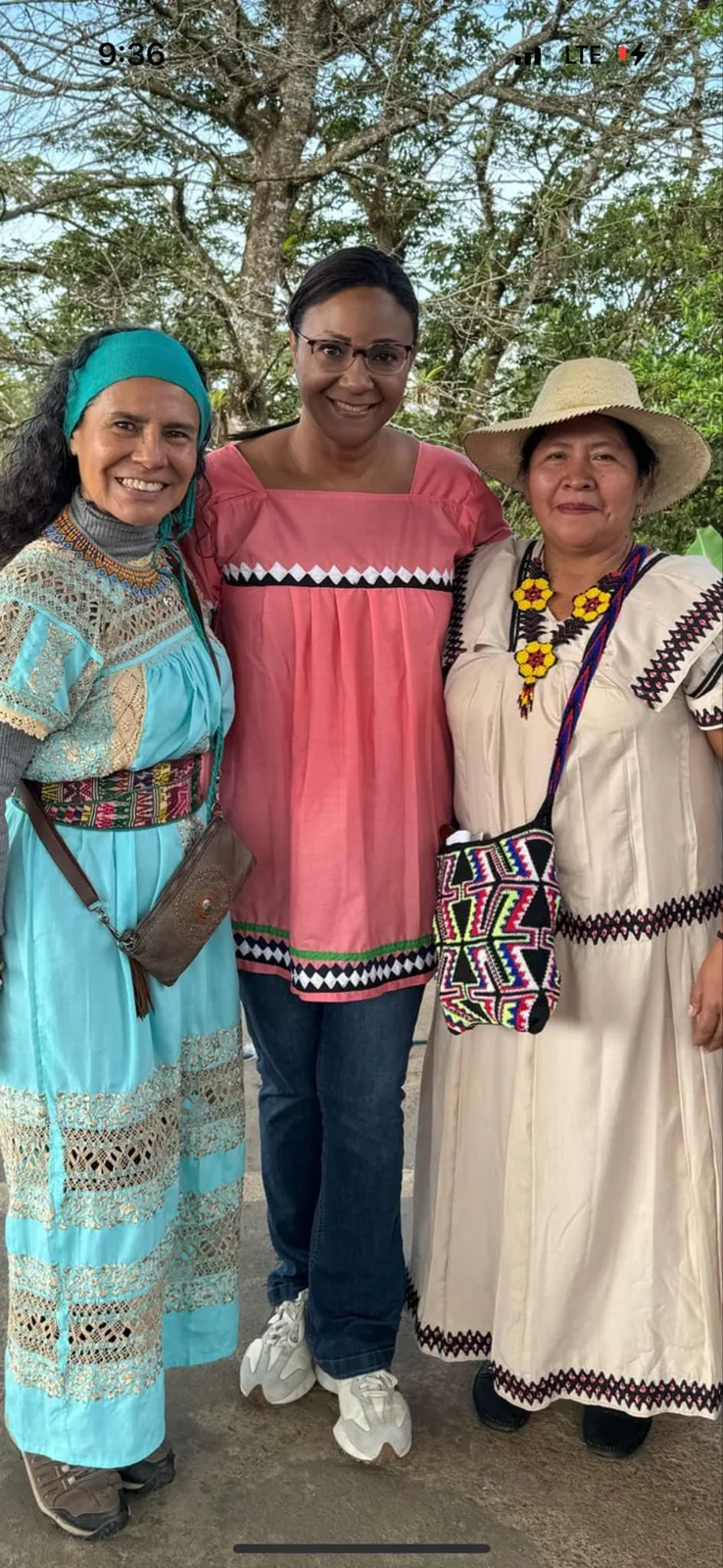 Three smiling women stand together under trees, one in a turquoise lace dress and headband, one in a pink embroidered blouse, and one in a cream dress and straw hat.