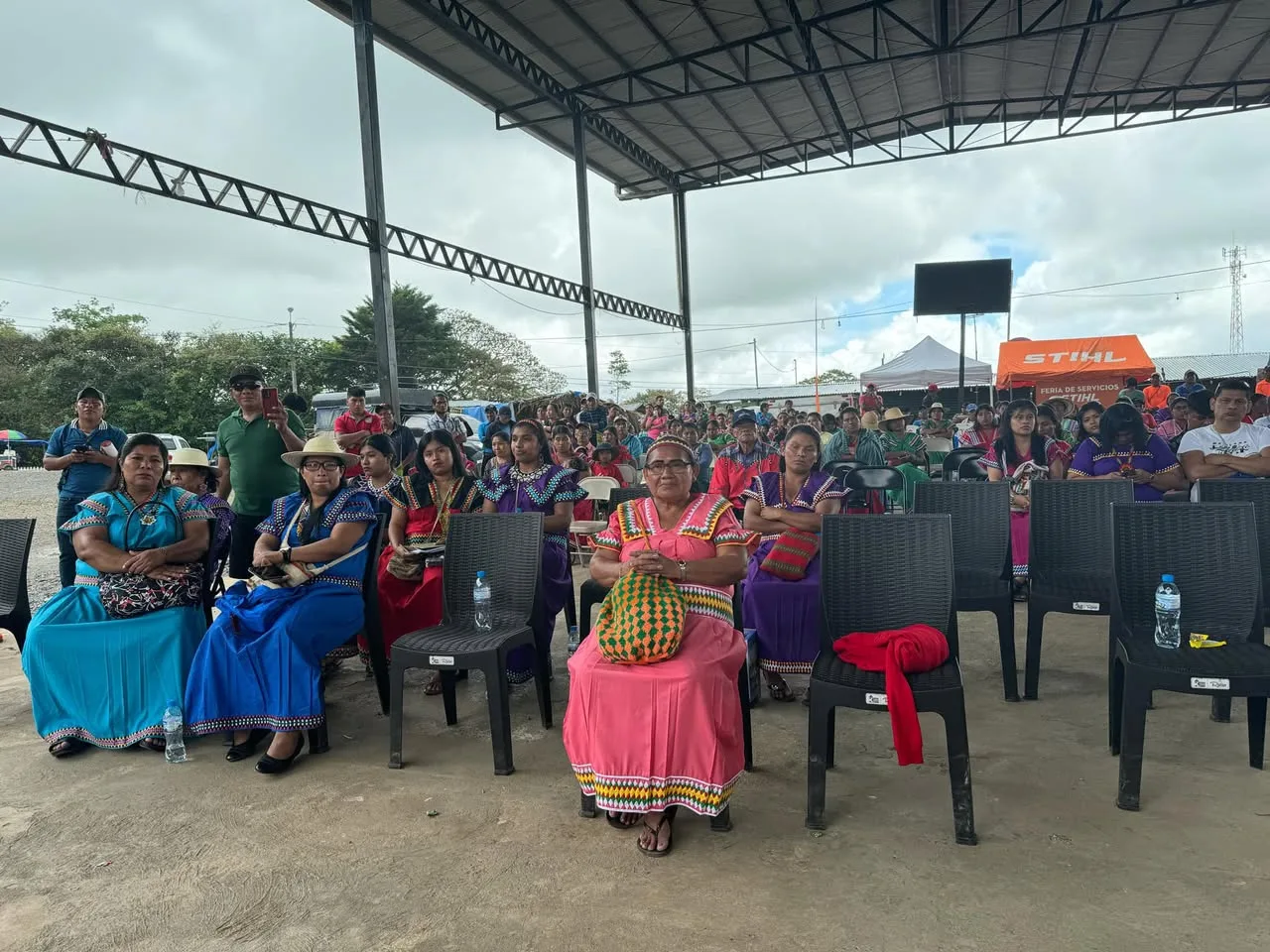 Indigenous women in brightly colored traditional dresses sit in chairs at the front of a large crowd gathered under an open metal-roofed pavilion.