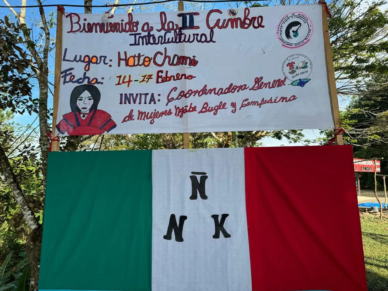 Hand-painted banner welcoming visitors to the II Intercultural Summit in Hato Chami, February 14-17, hosted by the General Coordinator of Ngäbe-Buglé and Campesino Women, above a green, white and red flag.