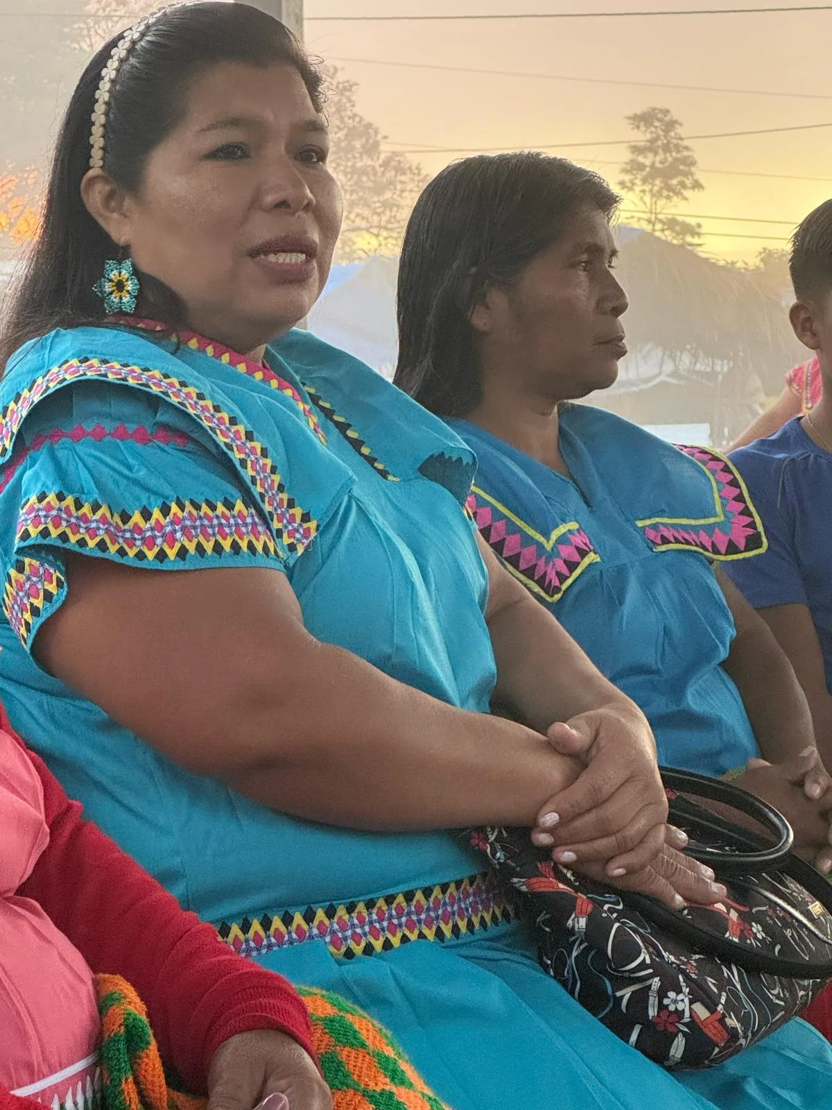 Two Ngäbe women in turquoise traditional dresses with colorful geometric trim sit side by side, one speaking with hands clasped over a handbag.