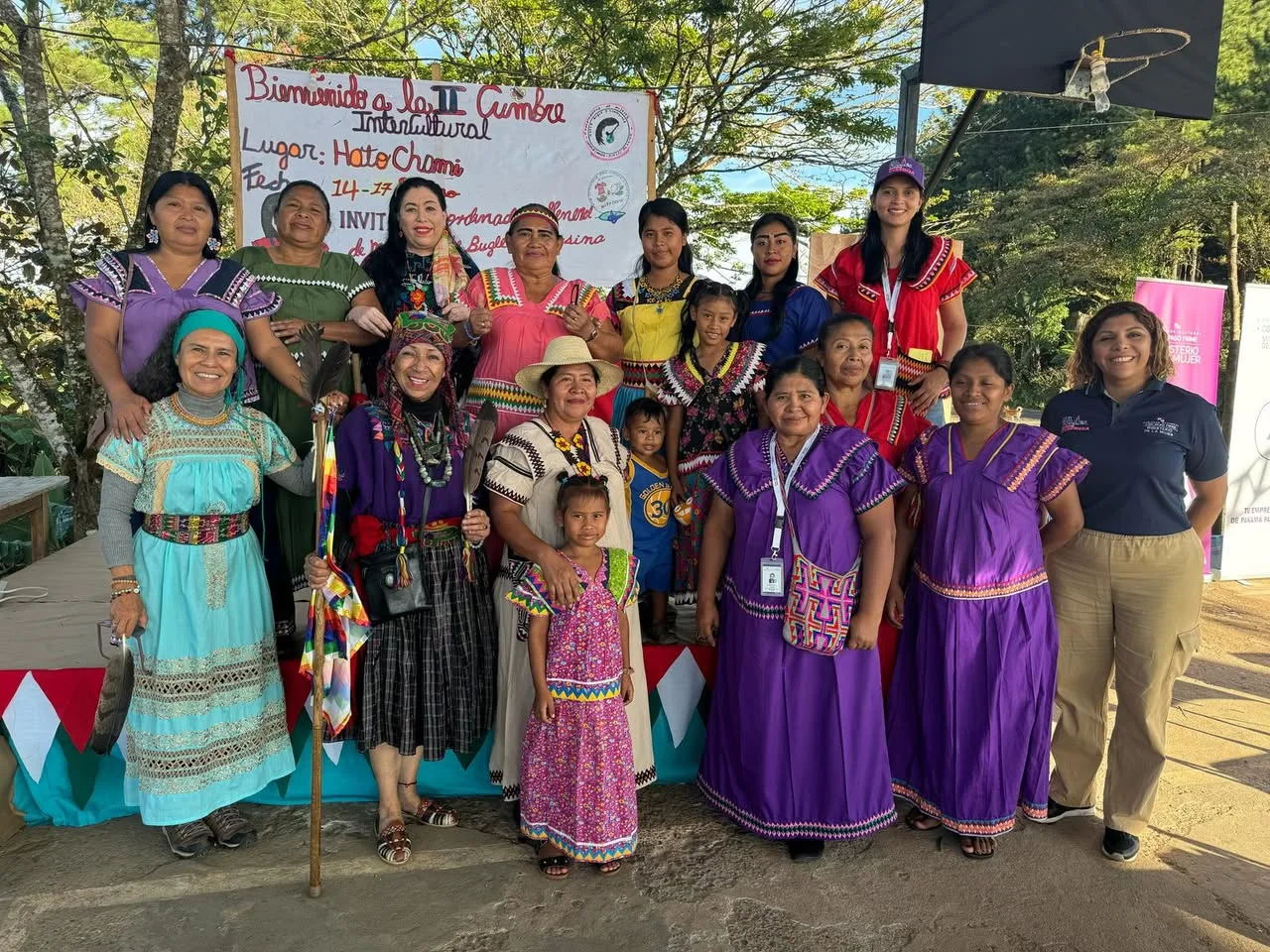 A large group of Indigenous women, children and allies in colorful traditional dress pose in front of the II Intercultural Summit banner, one holding a feathered staff.