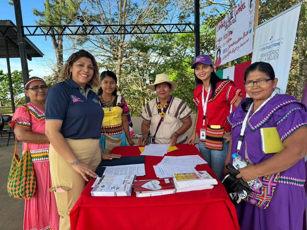 Six women, some in traditional dress and some in staff attire, smile around a red table with forms and pamphlets beside a Ministry of Women banner.