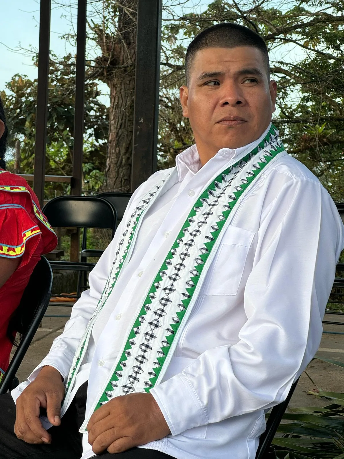 A man in a white shirt with green and black geometric trim sits on a folding chair, glancing to the side.
