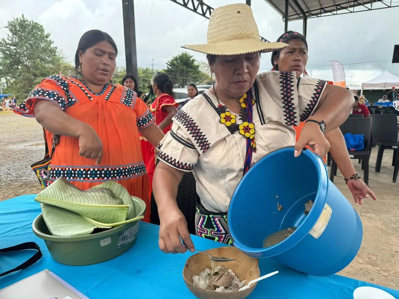 A woman in a straw hat serves soup over rice into a gourd bowl from a blue bucket while a woman in an orange dress looks on.