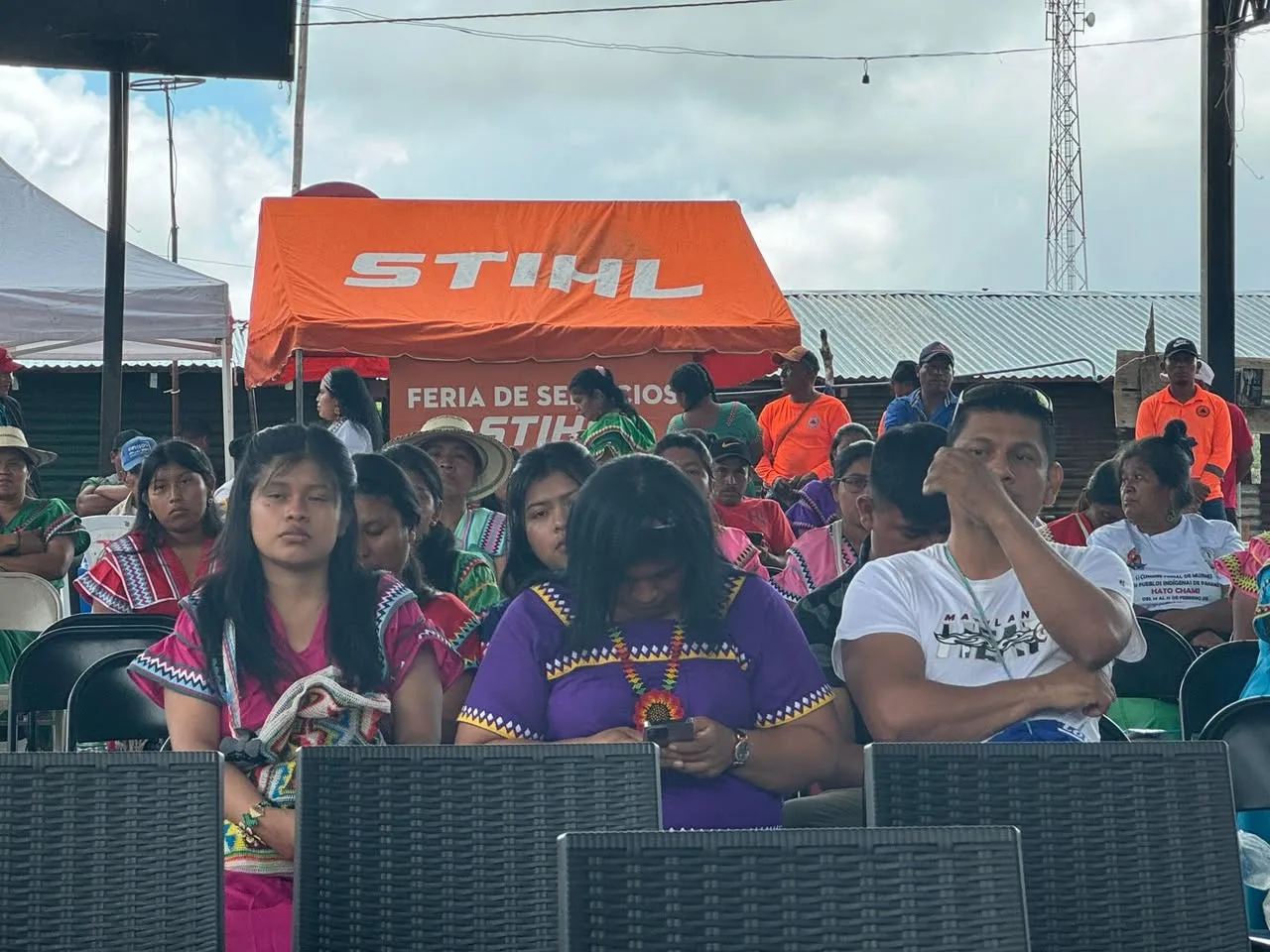 Participants in traditional dress and casual clothes sit under a pavilion, with an orange service tent and crowd behind them.