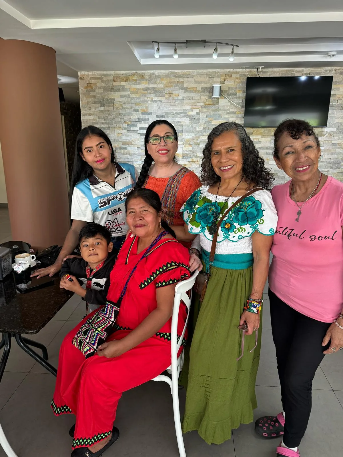 Six women and a young boy pose together indoors at a café table, several wearing traditional embroidered dresses.