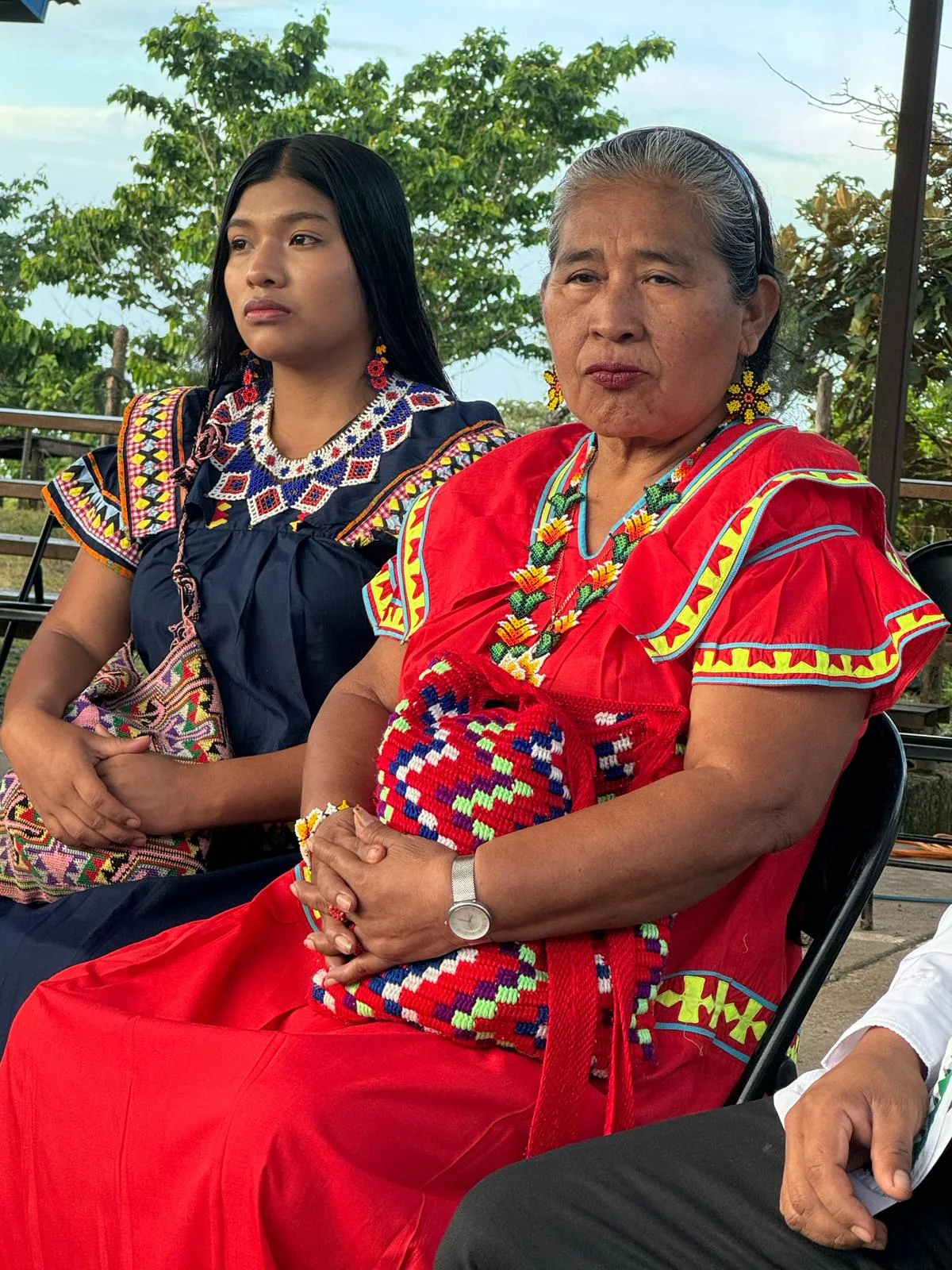 A young woman in a navy dress with a beaded collar and an elder in a red dress with a woven bag sit side by side.