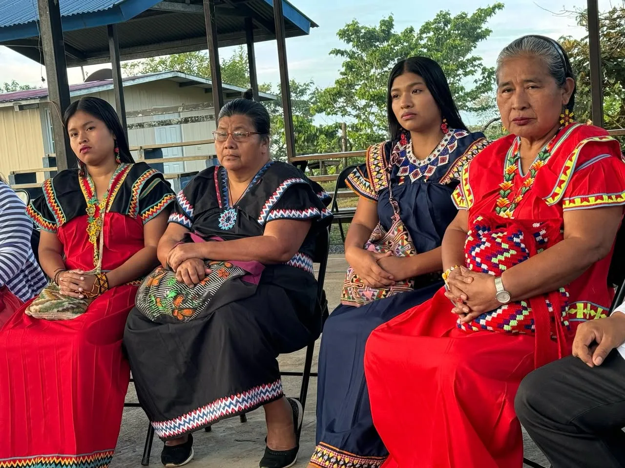 Four Indigenous women of different ages in red, black and navy traditional dresses with beaded collars sit in a row beneath a shelter.