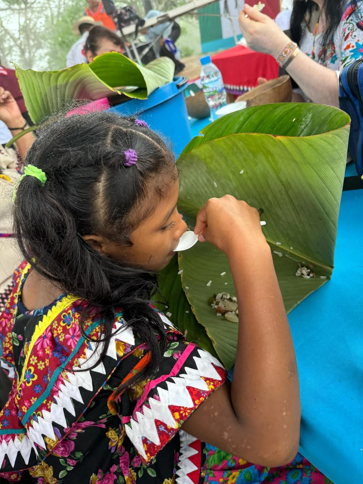A young girl in a black floral dress with white zigzag trim eats with a spoon from a large banana leaf at a blue table.