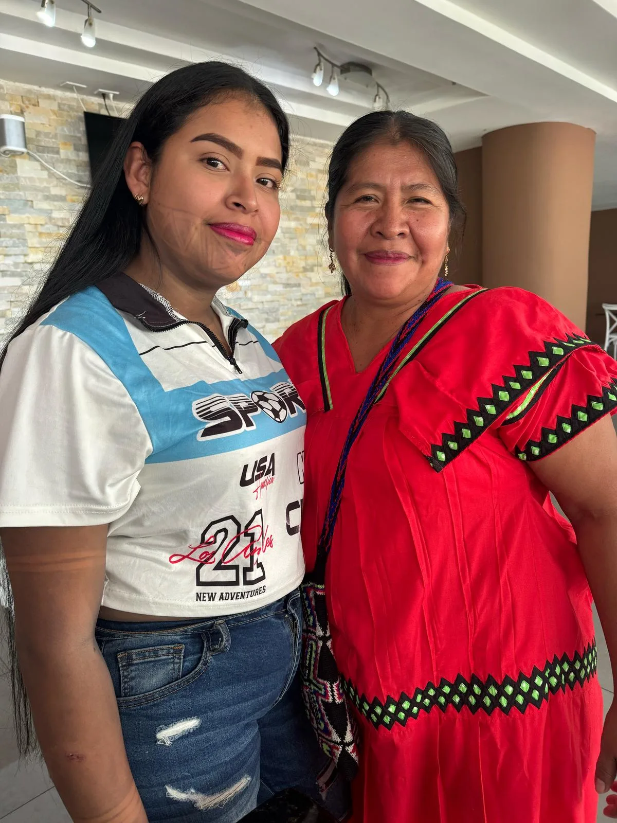 A young woman in a white sports shirt and an older woman in a red traditional dress stand close, smiling.