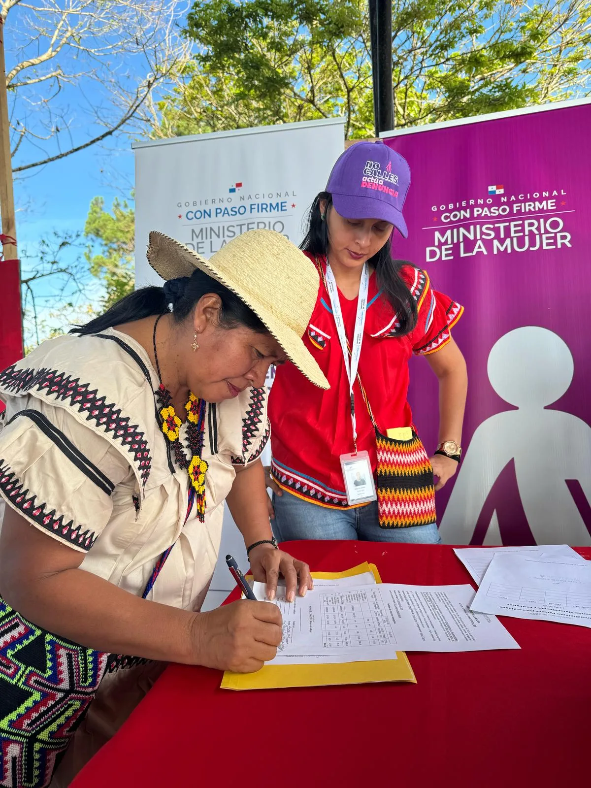 A woman in a straw hat signs a document at a red table as a staff member in a purple cap watches, beside Ministry of Women banners.