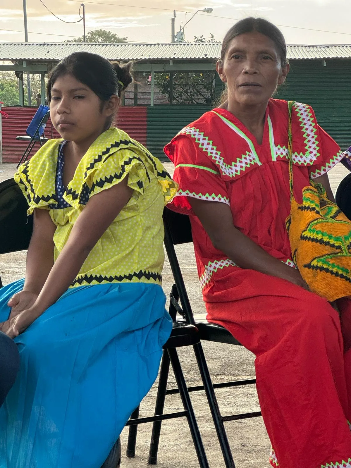 A young girl in a yellow and blue dress and a woman in a red dress with zigzag trim sit on folding chairs in an open courtyard.