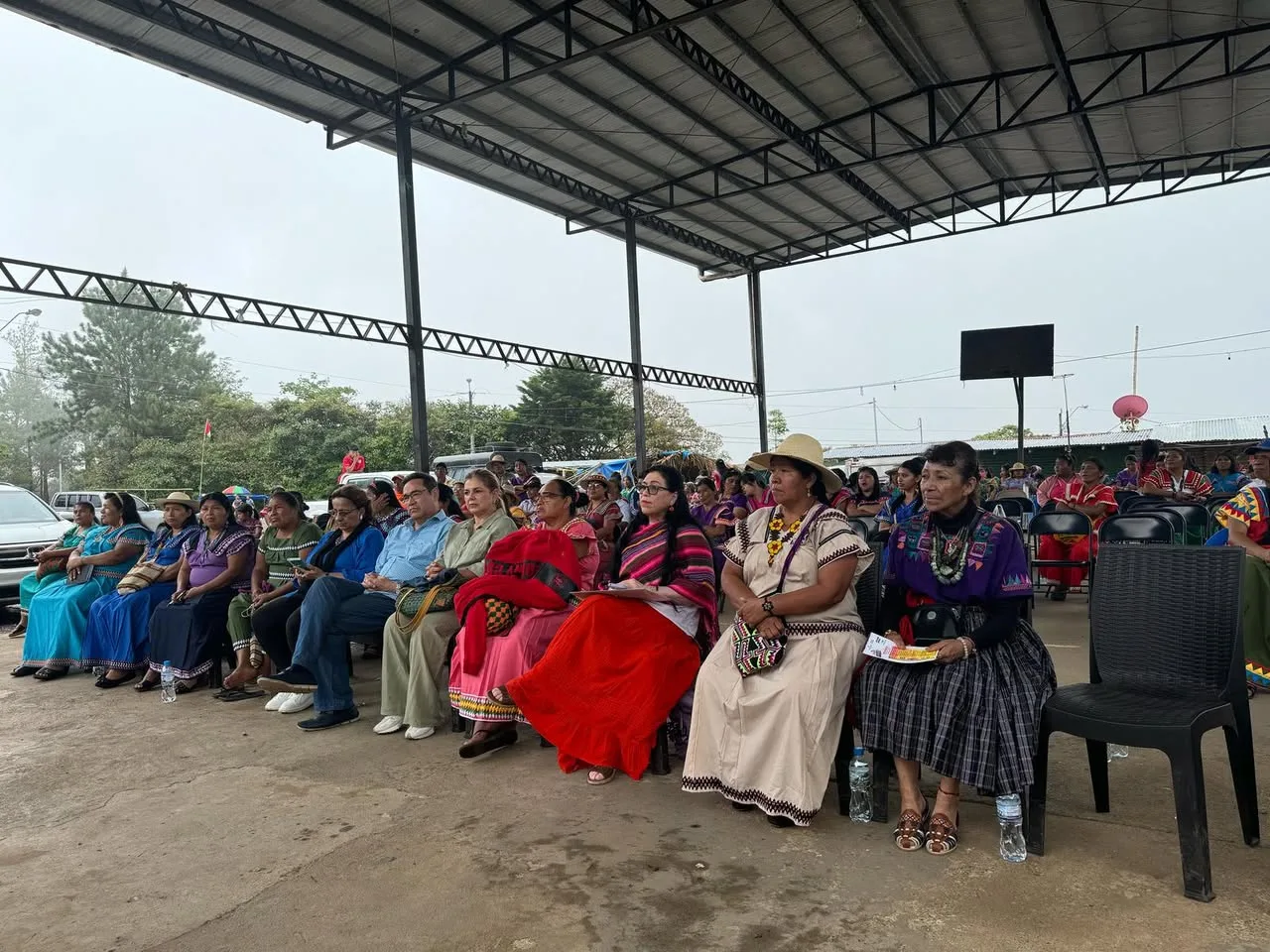 A row of women in traditional and everyday dress sit under a large metal-roofed pavilion, with a crowd of participants behind them.