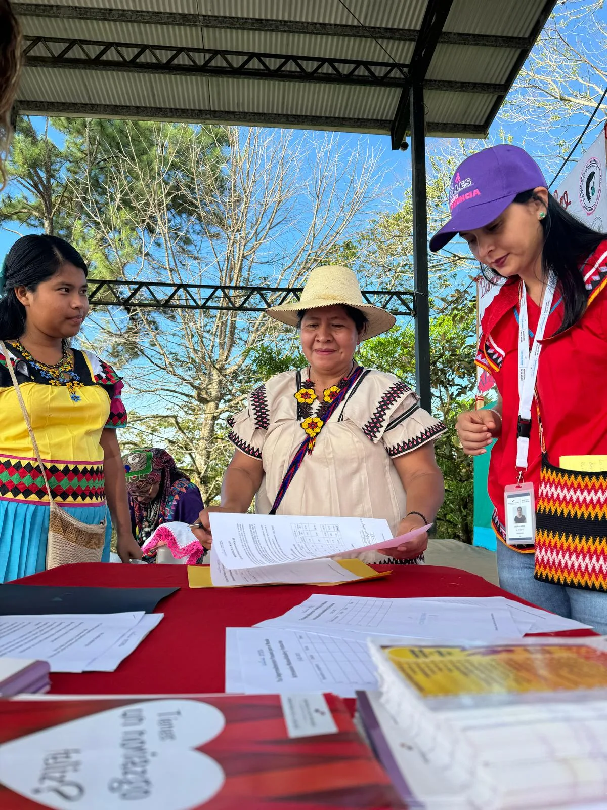 A woman in a straw hat and cream embroidered dress reviews papers at a red-covered table, joined by a young woman in yellow and a staff member in a purple cap.
