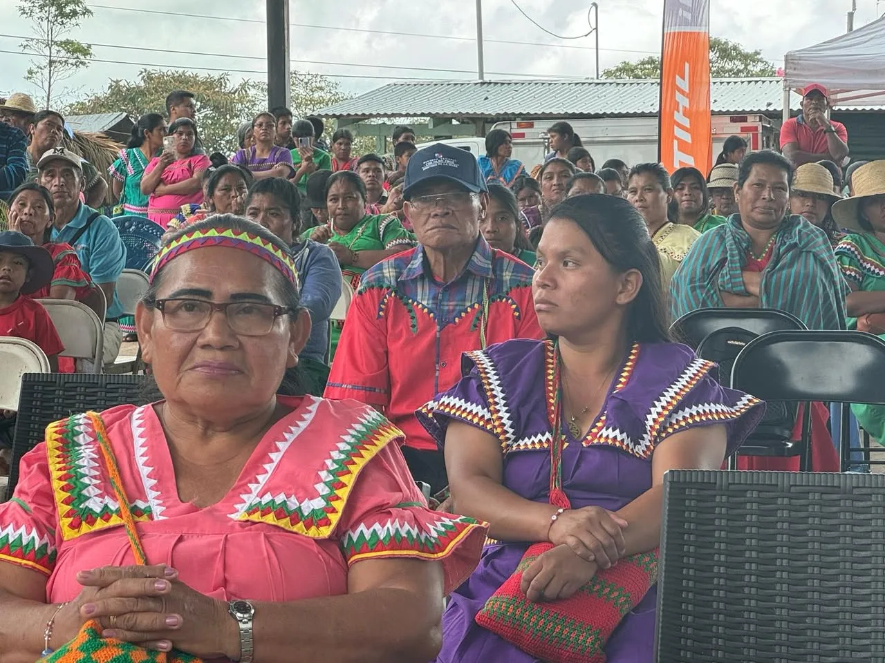 Older and younger women in pink and purple traditional dresses sit attentively at the front of a large crowd at the gathering.