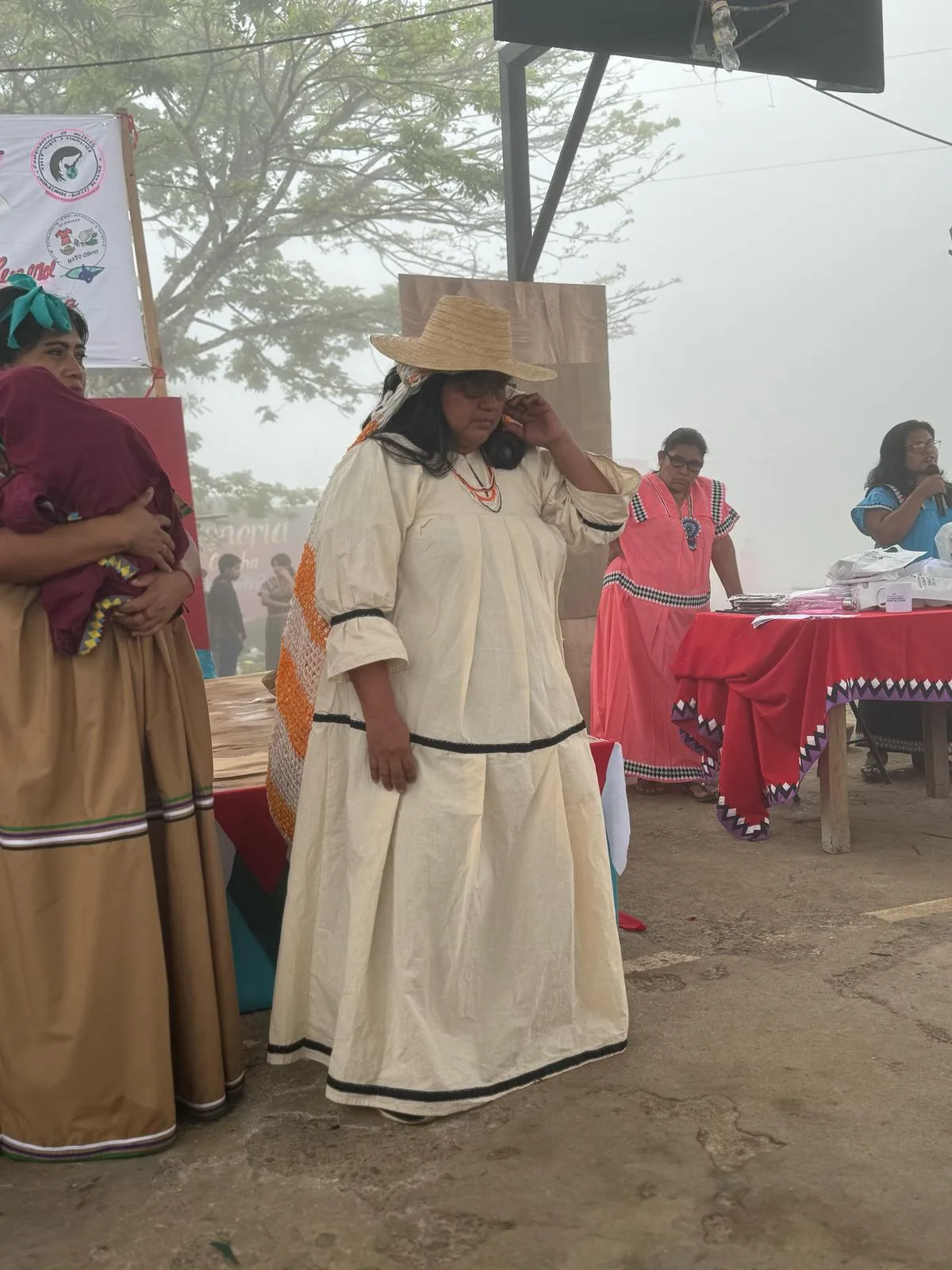 A woman in a cream dress and straw hat stands on a misty courtyard among other women in traditional dress near a table and banner.
