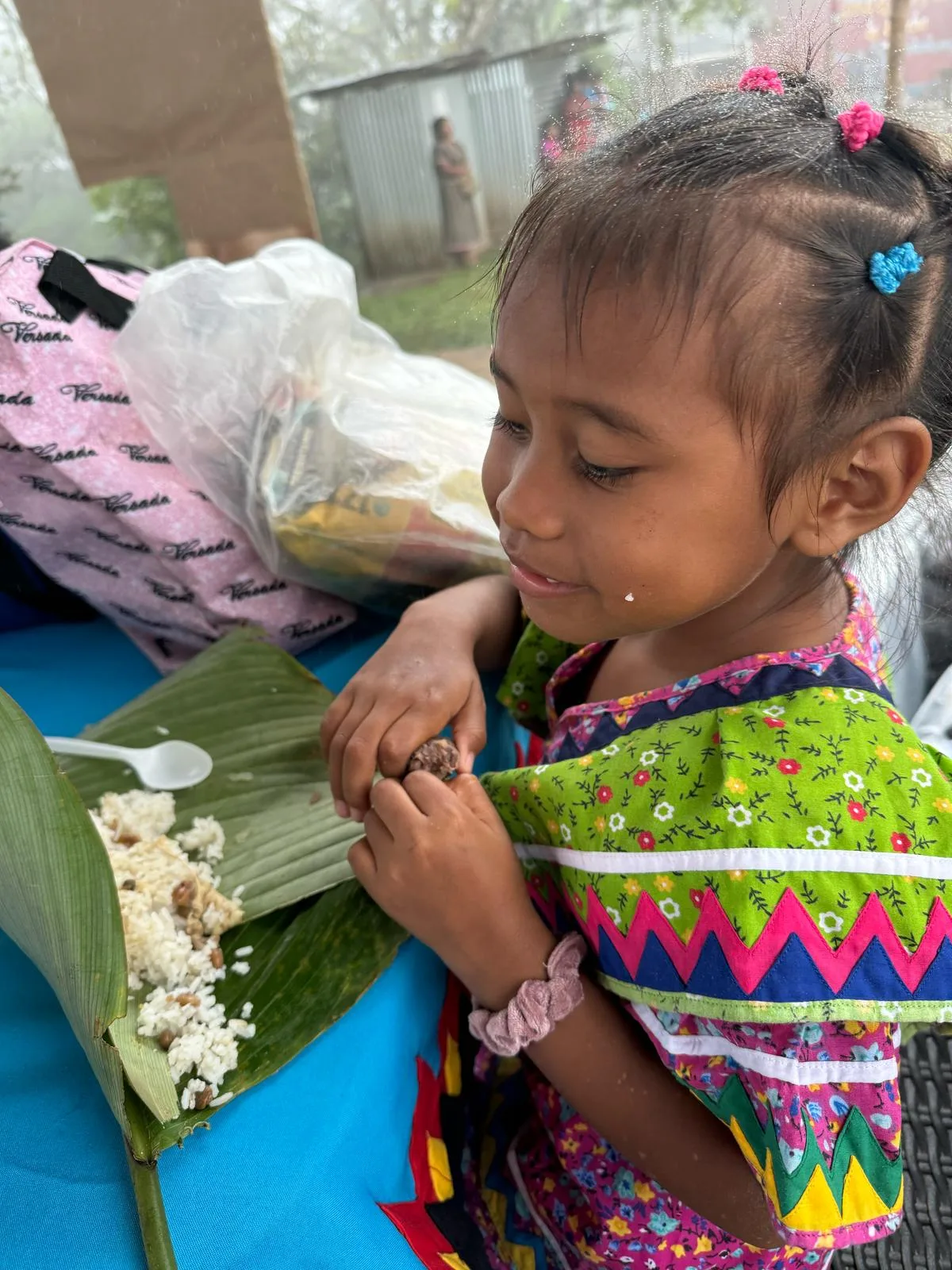 A little girl in a green floral dress with zigzag trim eats rice from a banana leaf at a blue table.